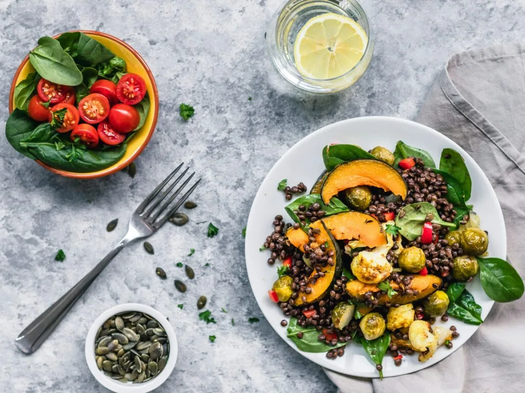 Teller mit Belugalinsen, Kürbis, Rosenkohl und Spinat, daneben Tomatensalat und Kürbiskerne
